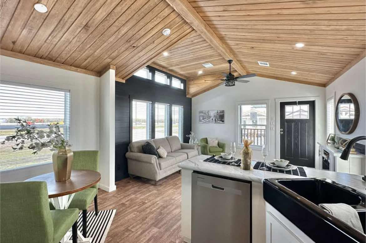 Modern living room with wood-paneled ceiling, large windows, beige sofa, green chairs, and a small dining table. Bright and inviting atmosphere.