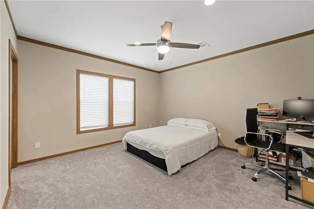 Bedroom with beige walls and carpet, featuring a ceiling fan, simple bed with white bedding, large window, and a tidy desk setup with a computer. Cozy and organized.