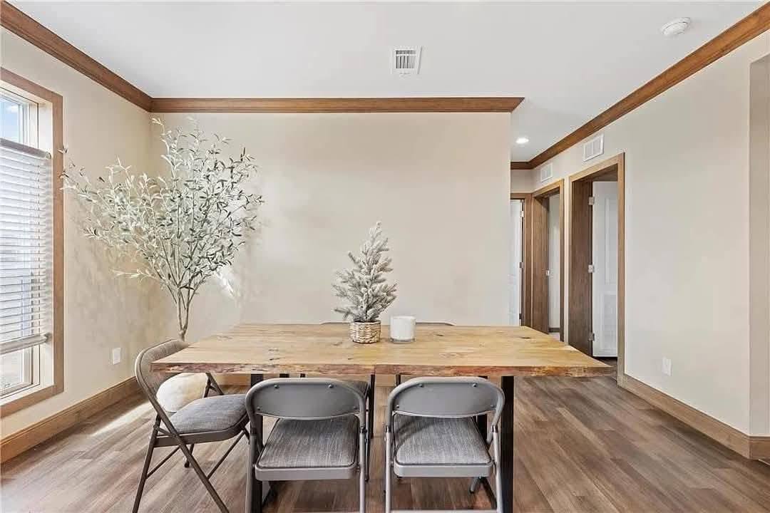 Modern dining room with wooden table and four chairs, decorated with a small plant centerpiece. A large potted plant stands by a sunlit window. Minimalist and inviting.
