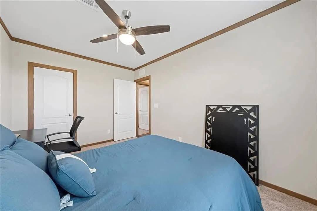 Bedroom with beige walls, blue bedding, and wooden trim. A ceiling fan hangs above, and a closed door and desk chair are in the background. Cozy ambiance.