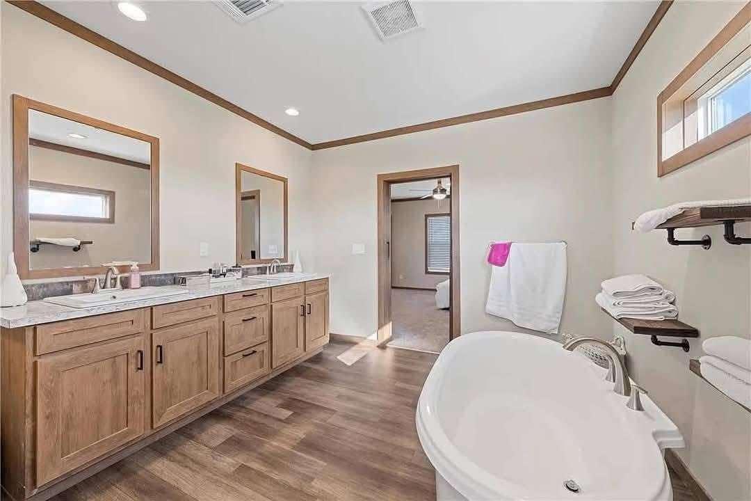 Spacious bathroom with wooden floor and cabinets, featuring a double sink vanity, freestanding tub, and towels on shelves. Bright, airy feel with natural light.