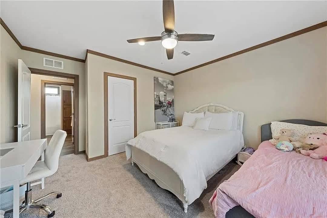 Bedroom with beige walls and carpet, featuring a double bed with white linens and a child's bed with pink covers and stuffed animals. There is a ceiling fan, a desk with an office chair, and wall art. The tone is cozy and inviting.