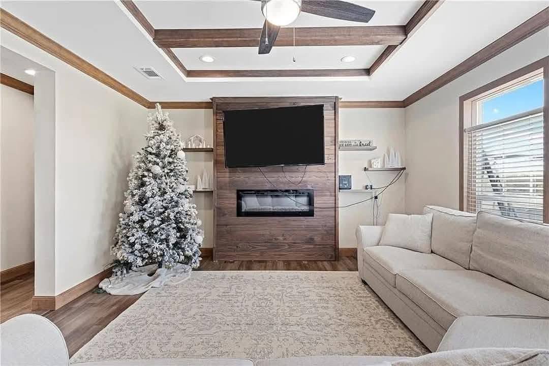 A cozy living room with a white, frosted Christmas tree, a beige sectional sofa, a large rug, and a wooden fireplace mantel with a TV above.