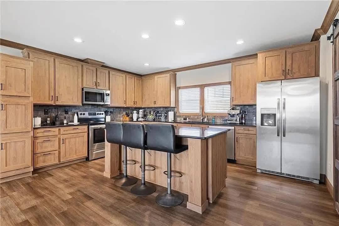 Bright kitchen with wooden cabinets and flooring, a central island with two black bar stools, stainless steel appliances, and recessed lighting. Cozy and modern.