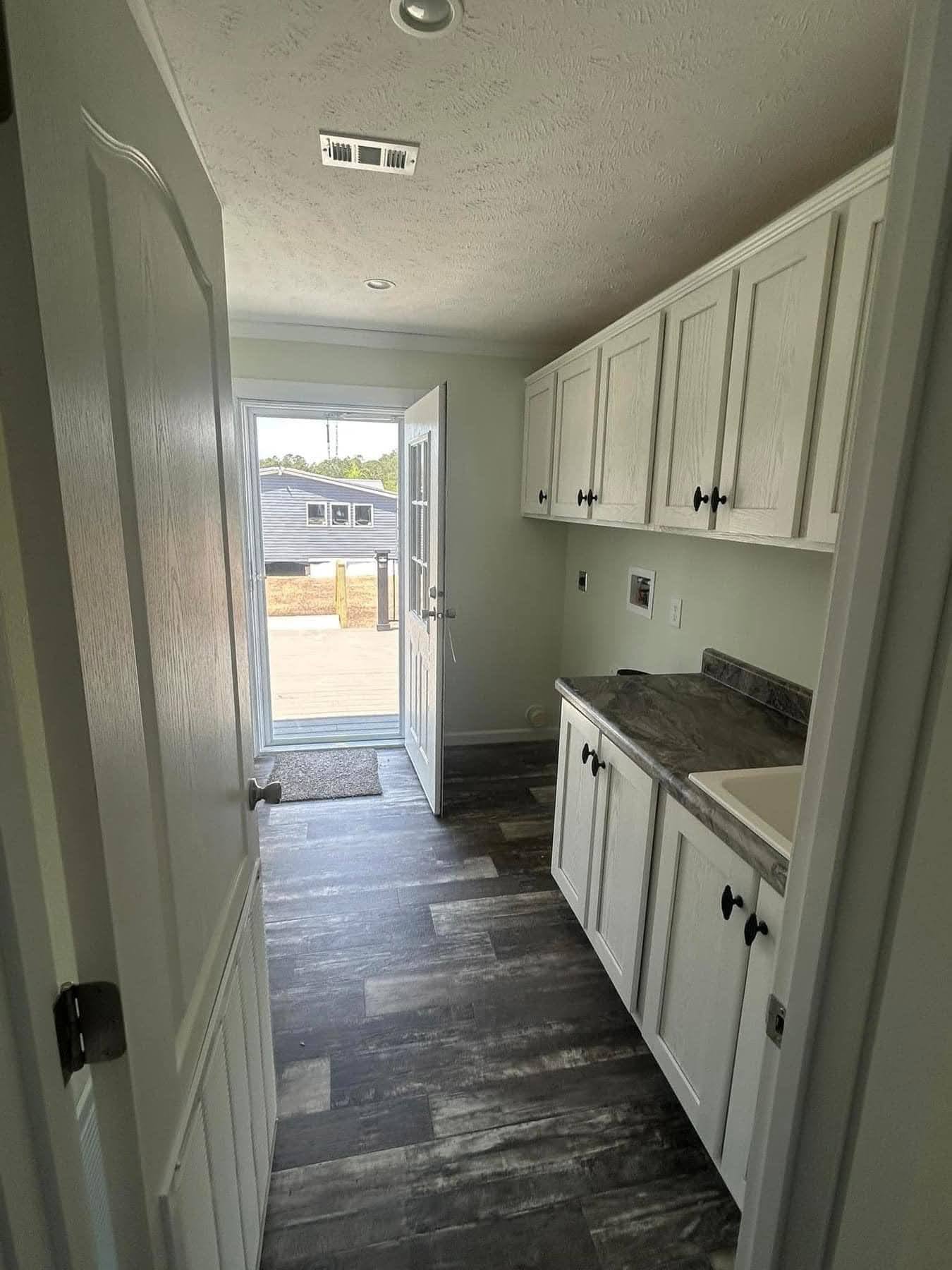 Narrow laundry room with white cabinets and a dark countertop on the right. Open door leads to outside, revealing sunny yard and distant house.