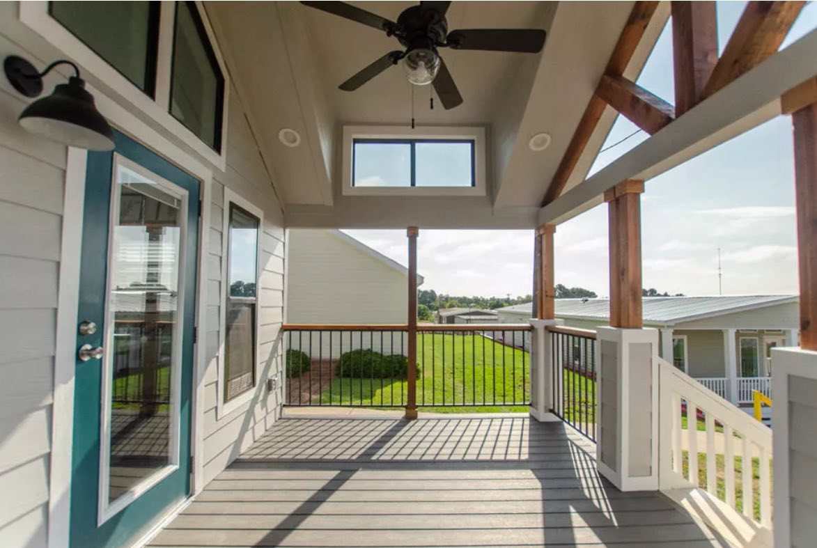 A sunlit porch with a ceiling fan, wooden beams, and railing overlooks a green lawn. The blue door and large windows create an airy, inviting space.
