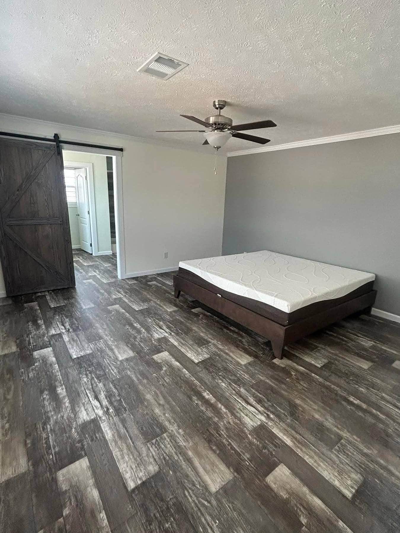 Spacious bedroom with gray walls, a dark wooden bed frame, and mattress on a textured wooden floor. A ceiling fan and sliding barn door enhance the modern vibe.
