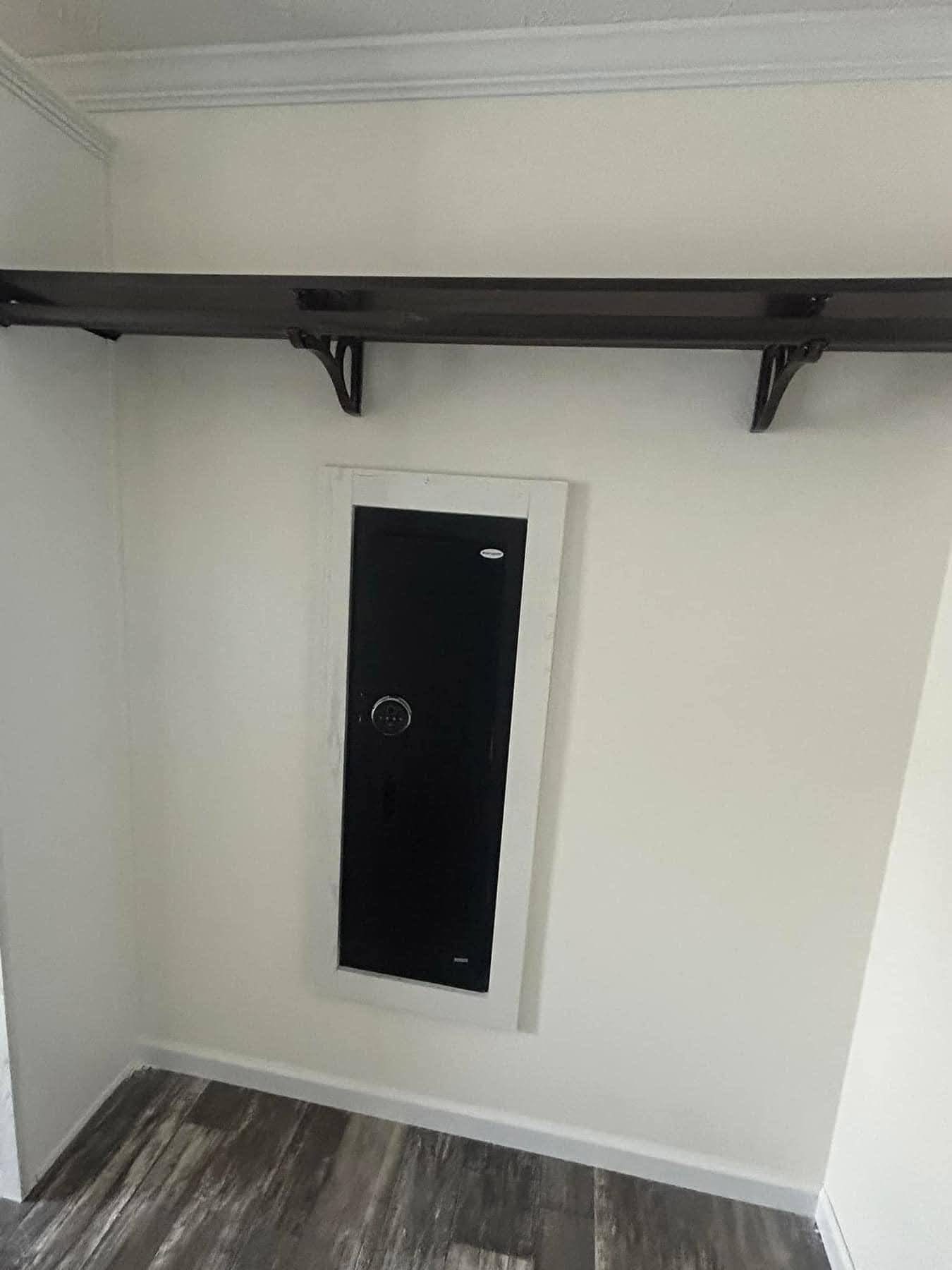 In a small closet, a black wall safe with a keypad is mounted vertically on a white wall below a dark wood shelf. The floor is wood-patterned laminate.