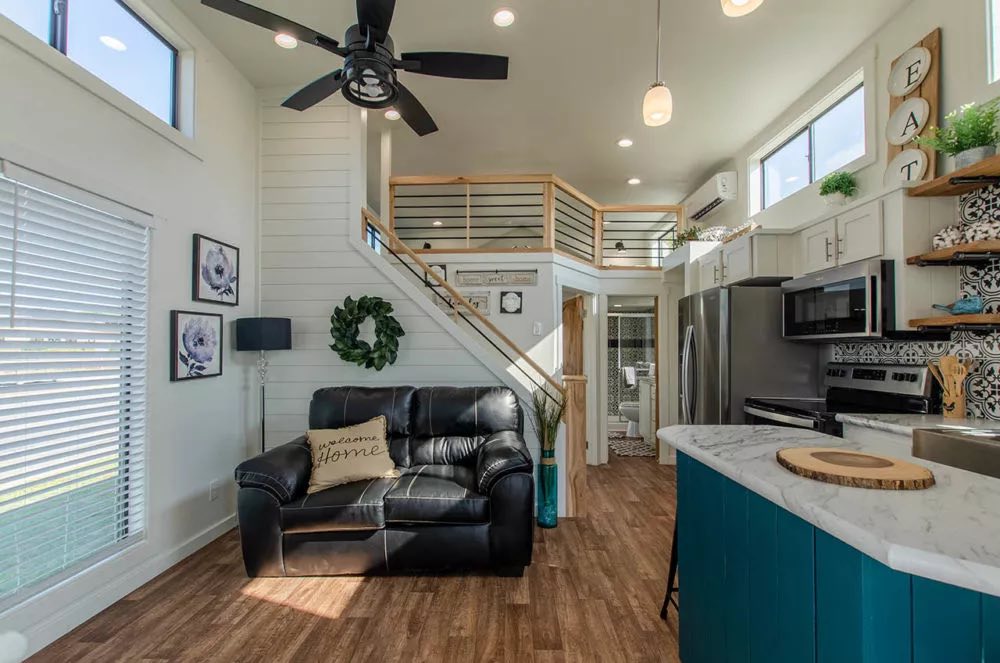 Modern tiny home interior with high ceiling and loft. Features a black leather couch, bright kitchen, wood accents, and decorative elements. Cozy ambiance.
