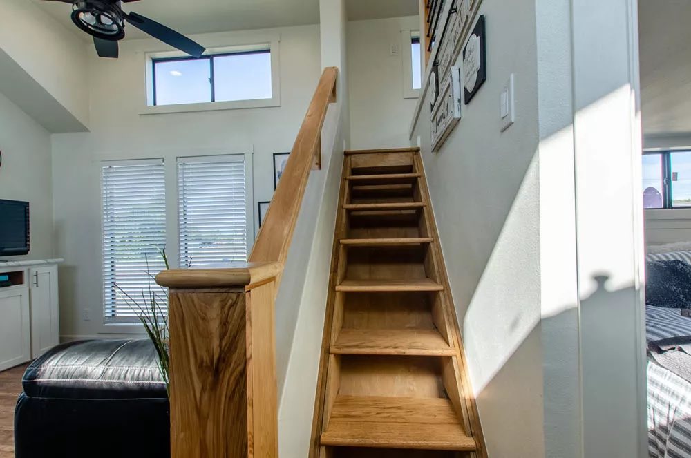 A cozy, sunlit interior showing narrow wooden stairs leading upstairs. A ceiling fan and window with blinds accompany a modern, minimalist style.