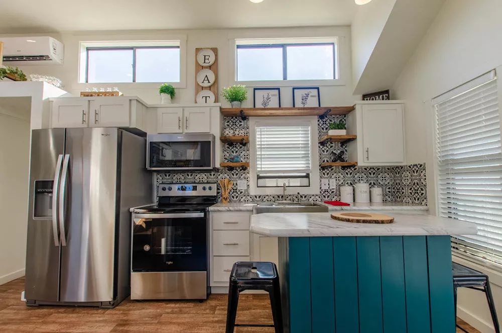 Modern kitchen with white cabinets, stainless steel appliances, and a teal island. Decorative tiles, plants, and "EAT" sign add warmth and style.