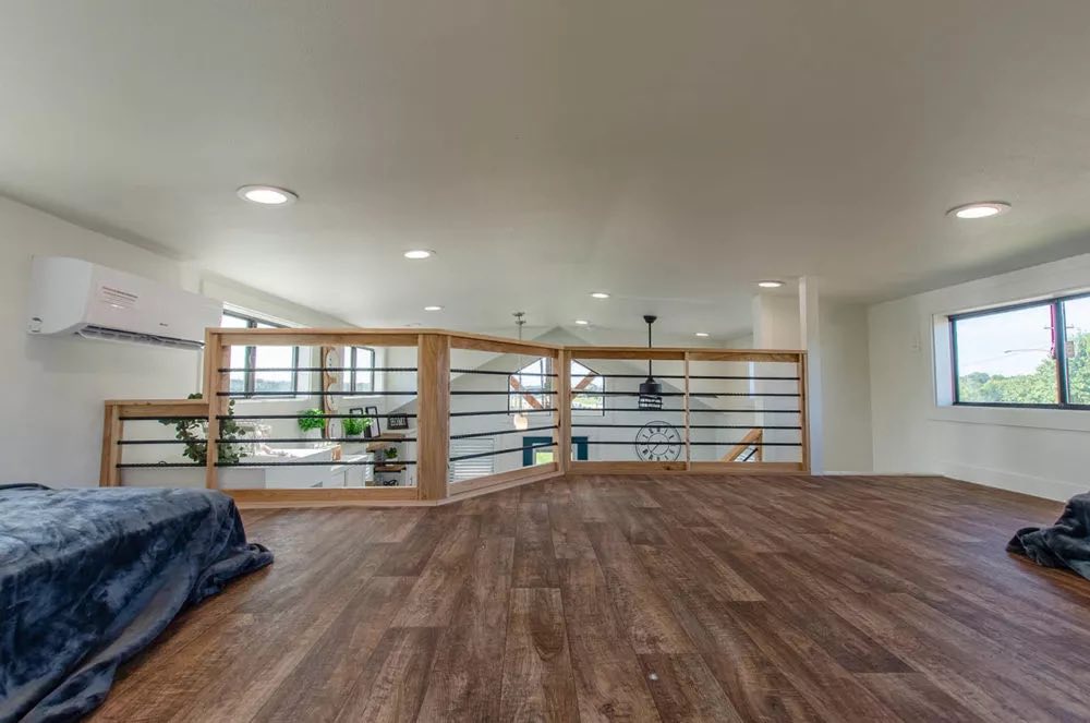 Spacious loft with wooden floors, minimal furniture, large windows, and railing overlooking the room below. Bright and airy atmosphere.