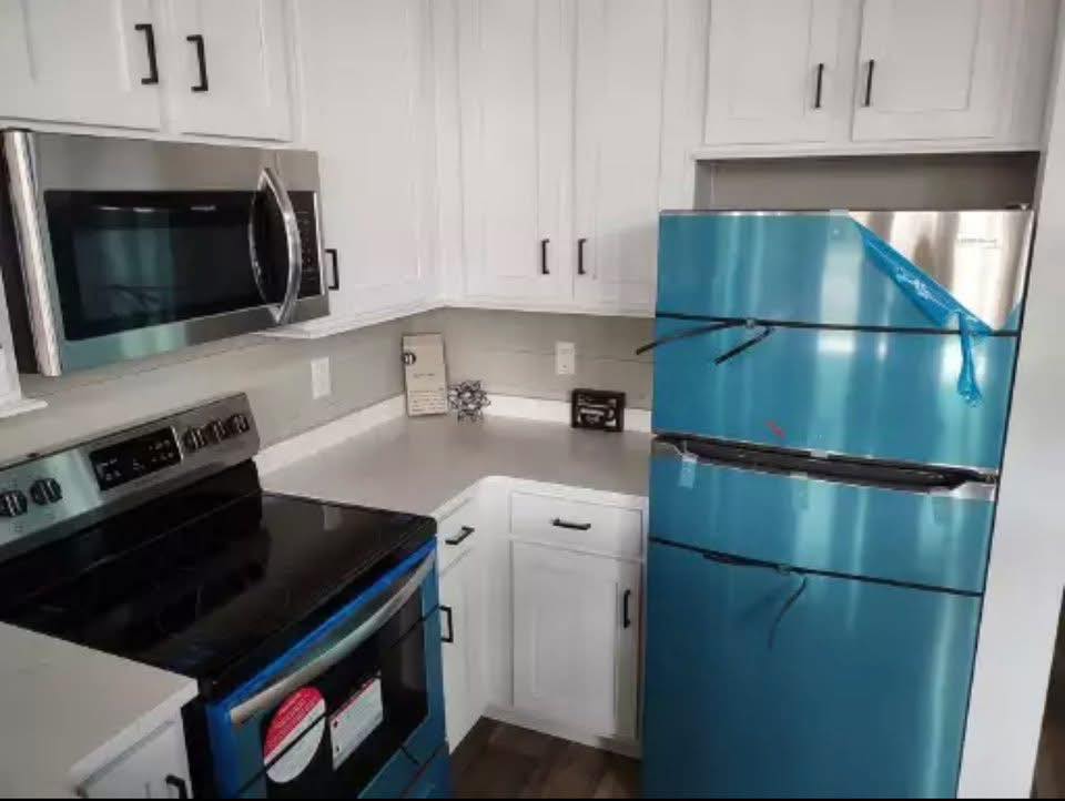 Modern kitchen with white cabinets, sleek stainless steel microwave and oven. A new fridge, wrapped in protective blue film, stands out. Clean, airy ambiance.