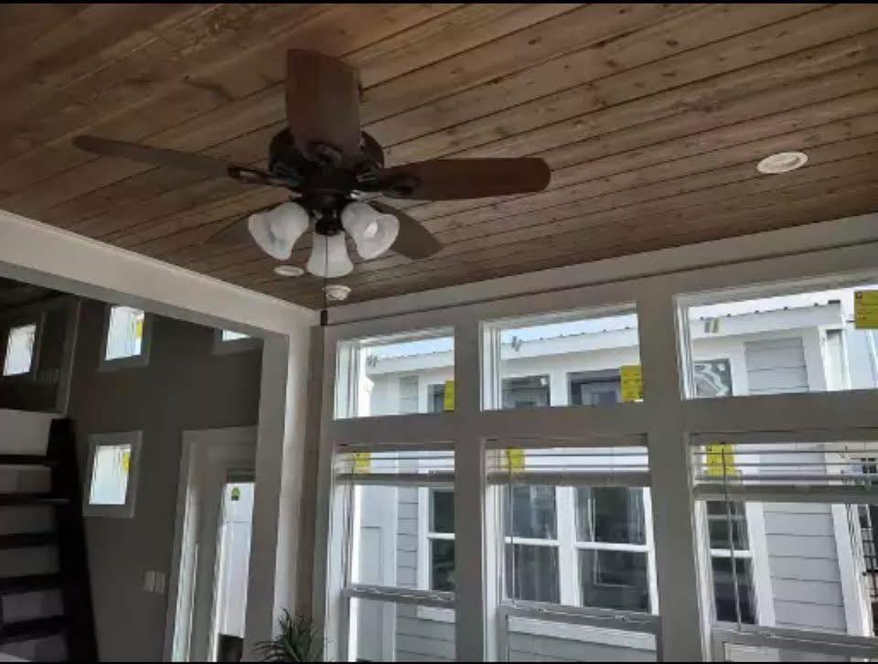 Ceiling fan with wooden blades and lights on a wood-paneled ceiling. Large windows with natural light create a bright, airy atmosphere in a modern room.