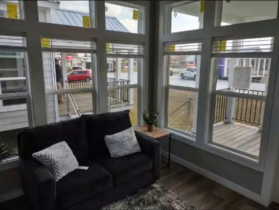 Cozy living room with large windows showcasing an outdoor view. A dark sofa with patterned cushions and a small table with a plant create a welcoming atmosphere.
