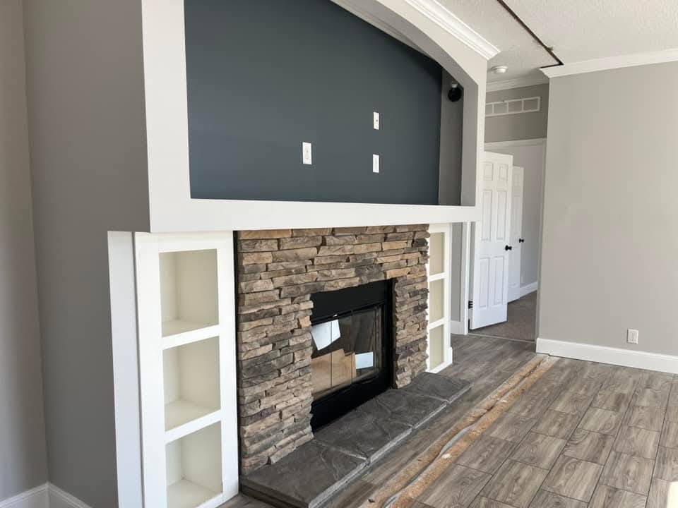Modern living room with a stone fireplace, white shelves, and dark blue accent wall. The room has wood flooring, creating a cozy atmosphere.