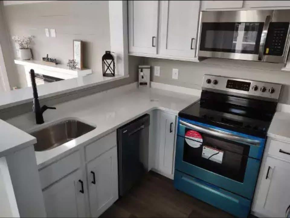 Modern kitchen with white cabinets, a shiny stainless steel microwave, and a blue oven. Minimalistic decor includes a black lantern and small plants.