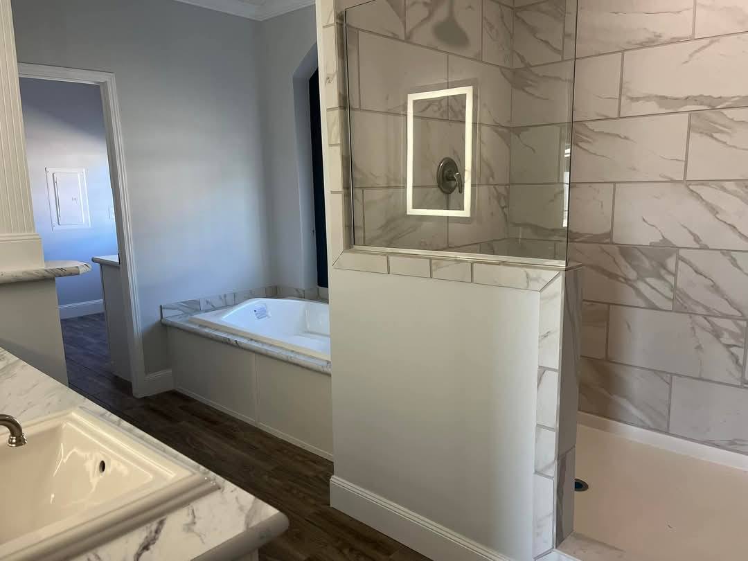 Elegant bathroom with a tiled shower featuring glass walls and a built-in recessed light. A white bathtub with marble accents is set against wooden floors.