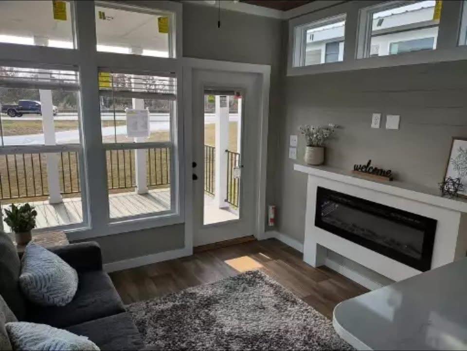Cozy living room with large windows and a glass door leading to a porch. Features a gray sofa, fireplace, welcome sign, and potted plants, exuding warmth.
