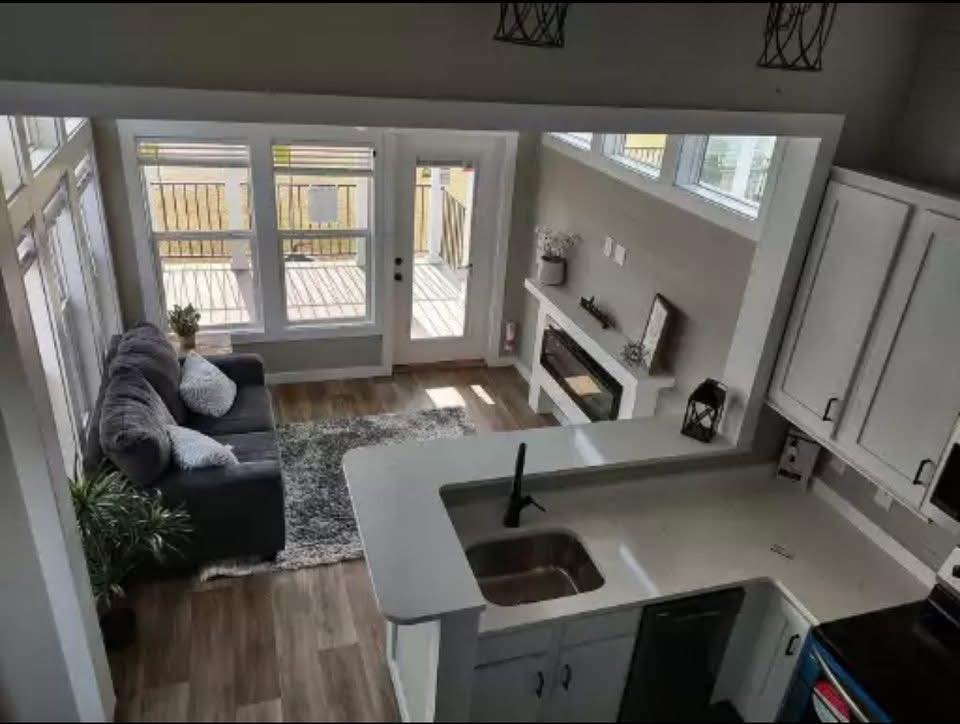 A cozy living room with a gray sofa, white pillows, and a small plant. A rug leads to a glass door, with sunlight streaming in. Kitchen counter with black faucet.
