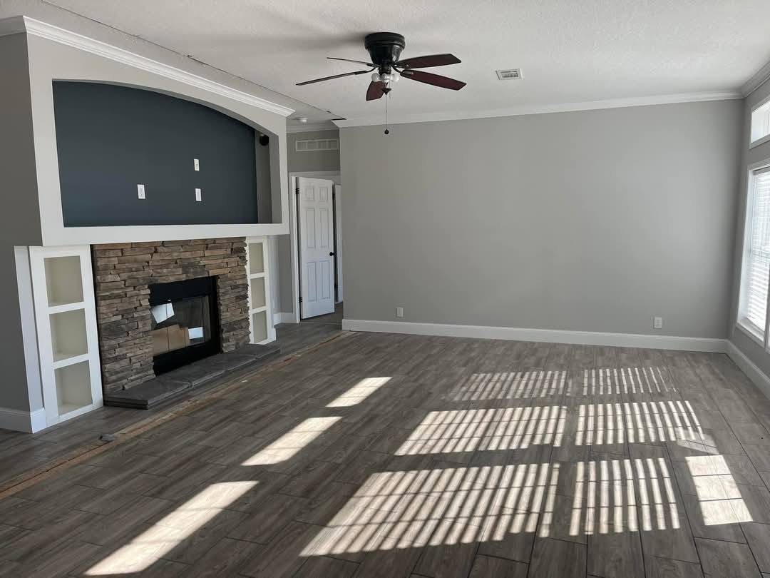 Spacious living room with gray walls, wood-like tile floor, and a stone fireplace. Sunlight creates window pane shadows. Cozy and modern feel.