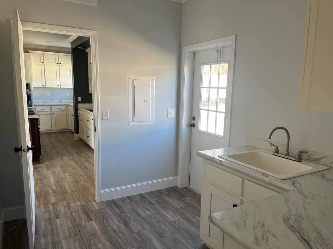 Bright kitchen with light gray walls, wood-style flooring, and white cabinets. The room features a marble countertop, sink, and a door with a window.