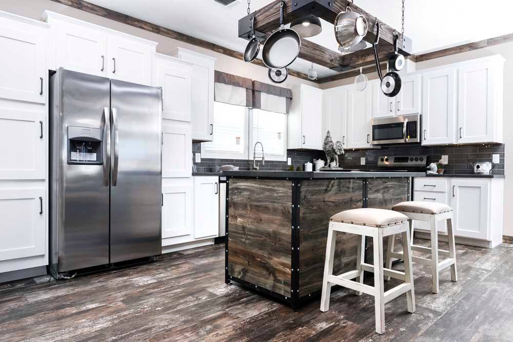 Modern kitchen with white cabinets, gray countertops, and a stainless steel refrigerator. A central island with wooden texture and two stools is beneath a hanging pot rack. The room has a rustic yet contemporary feel.