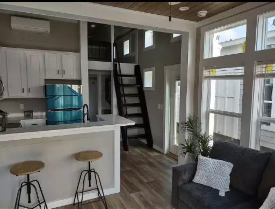 Modern tiny home interior with white cabinets, compact kitchen with bar and stools, dark ladder to loft, large windows, gray sofa, and cozy decor.