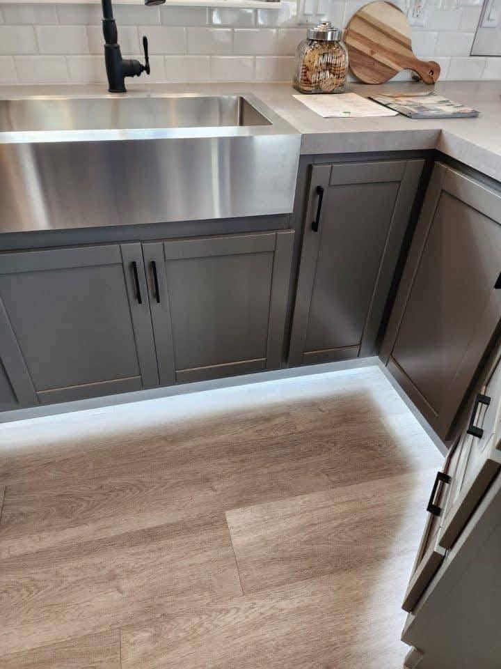 Modern kitchen with stainless steel farmhouse sink, dark gray cabinets, and black faucet. Light wood flooring contrasts subtly with white cabinets.