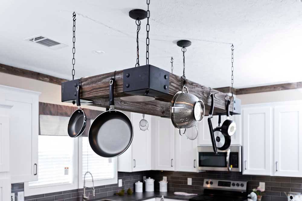 A rustic kitchen with a wooden pot rack hanging from the ceiling, holding various pans and kitchen utensils. White cabinets and stainless appliances create a cozy atmosphere.