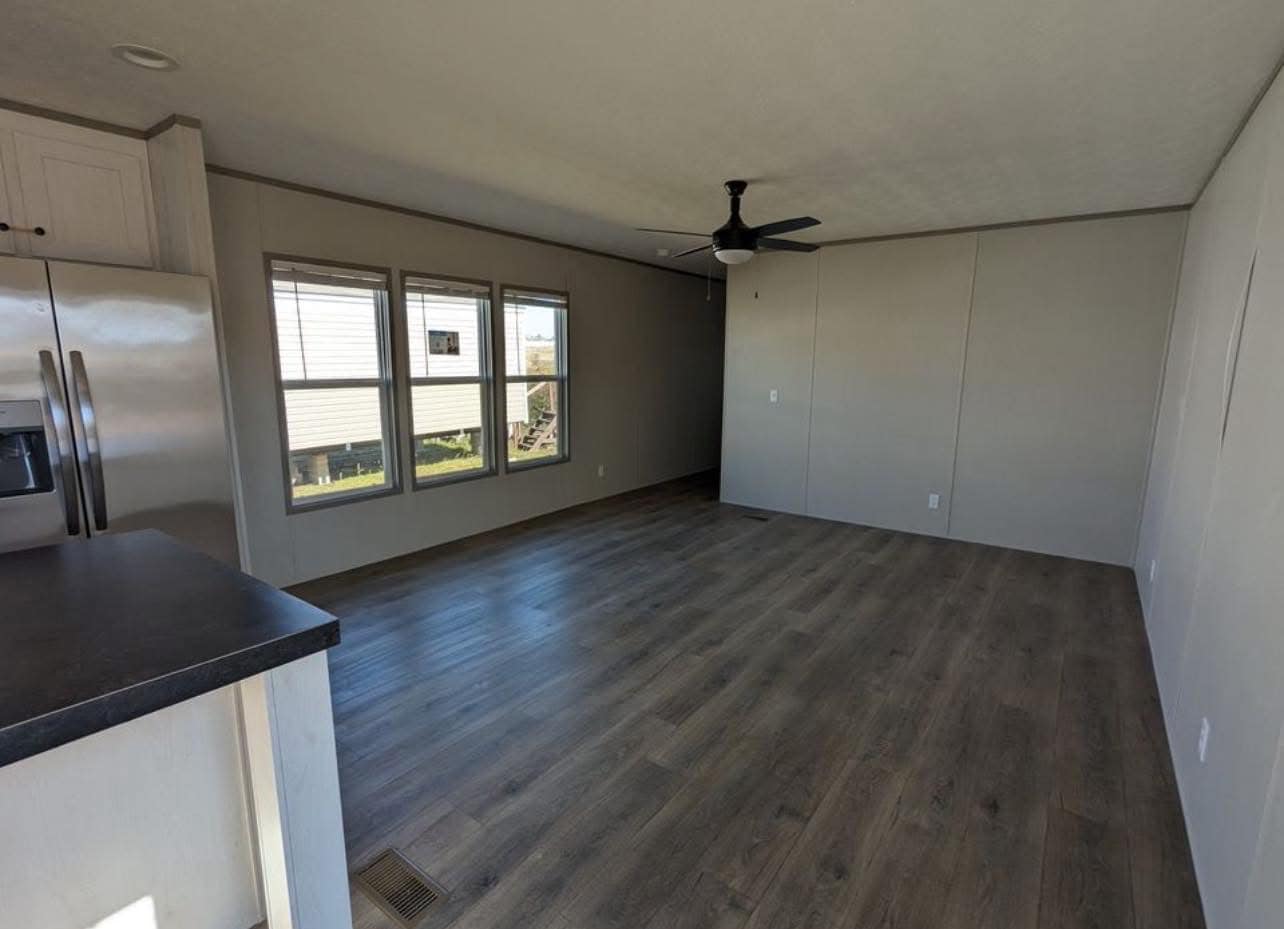 Spacious empty room with light wood flooring, three large windows, and a ceiling fan. The kitchen counter and stainless steel fridge are visible to the left.