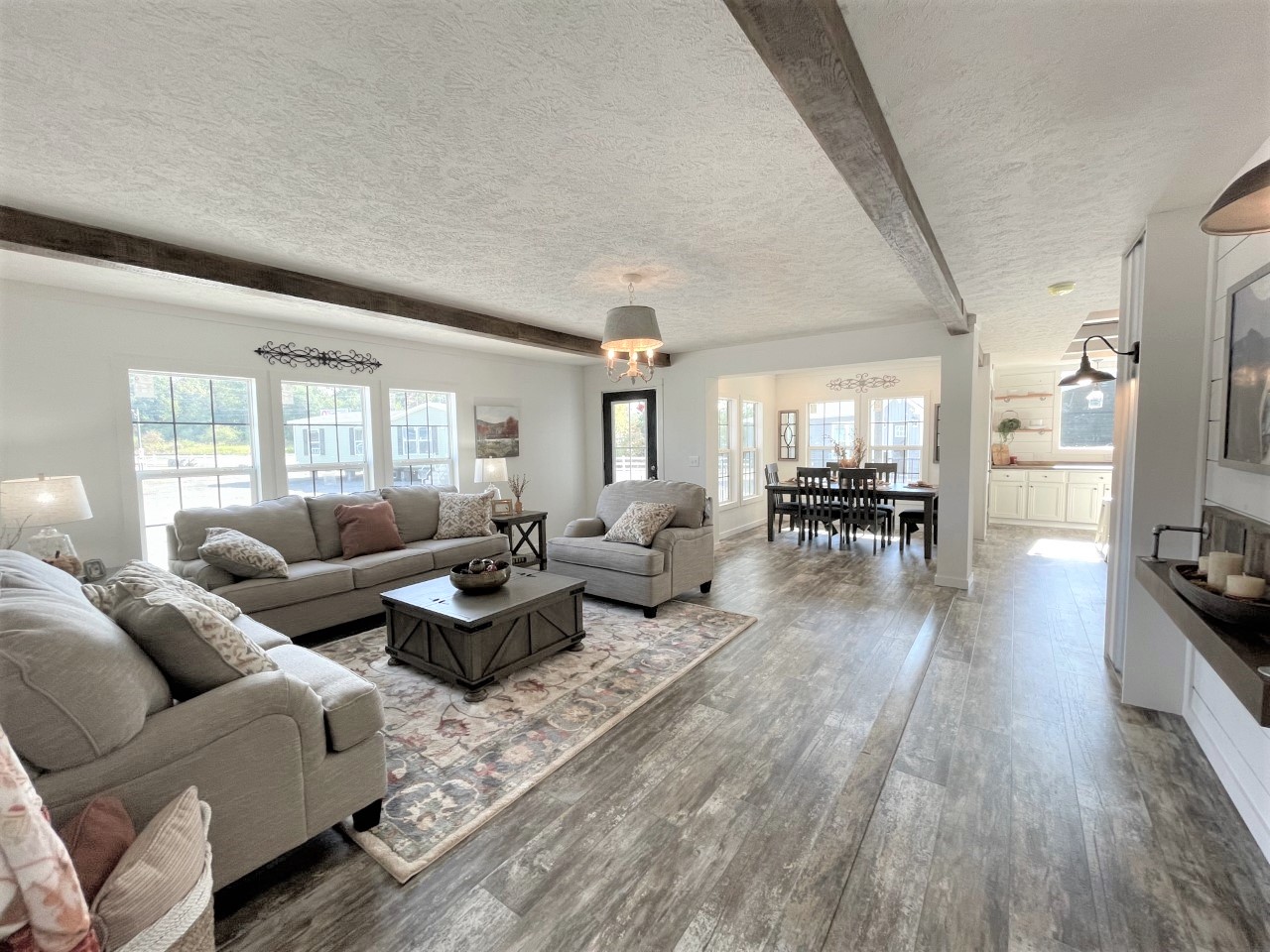 Spacious, well-lit living room with gray sofas, rustic wooden beams, a large dining table, and bright kitchen. Cozy and inviting atmosphere.