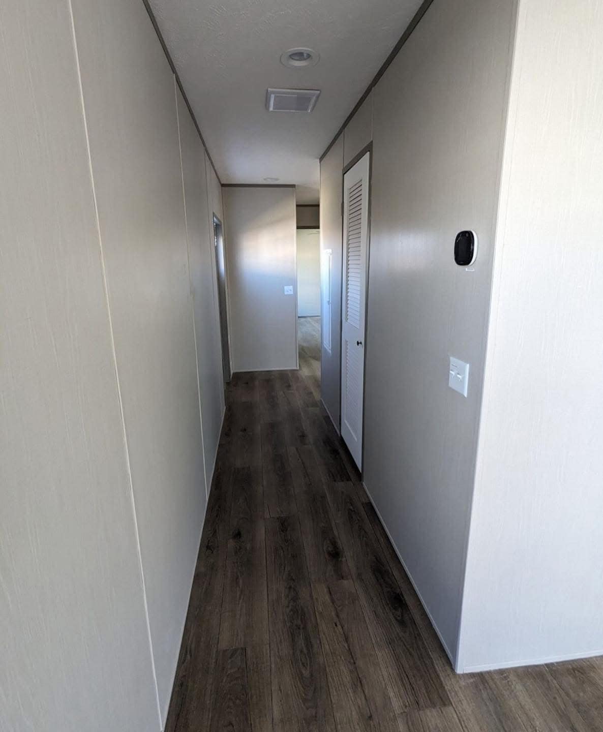 Narrow hallway with beige walls and dark wood flooring. A louvered door on the right, wall-mounted thermostat, and recessed lighting create a minimalist look.