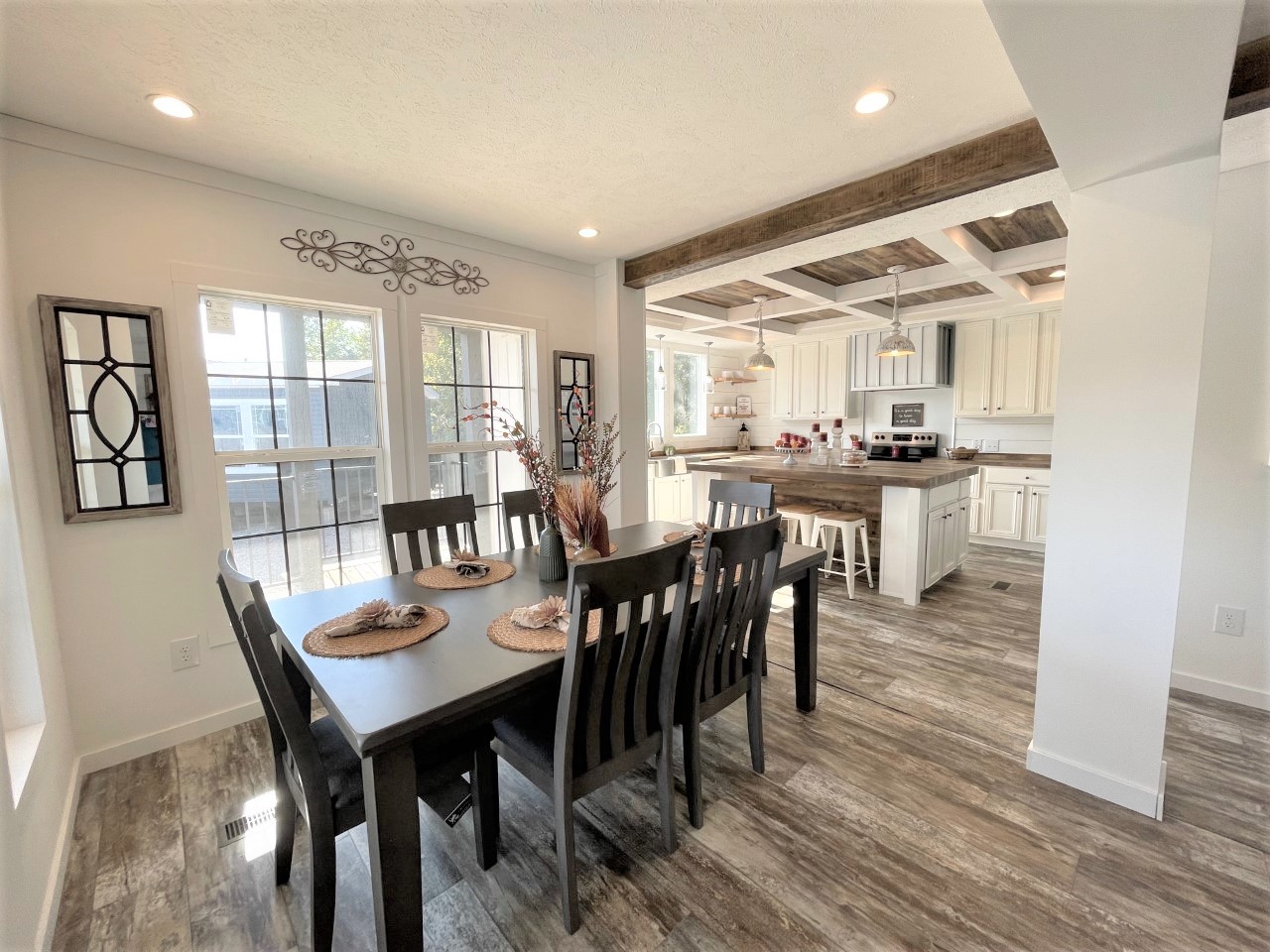 Bright dining room with a dark wooden table and chairs, adorned with natural centerpieces. Adjacent is a kitchen with white cabinets and rustic accents.
