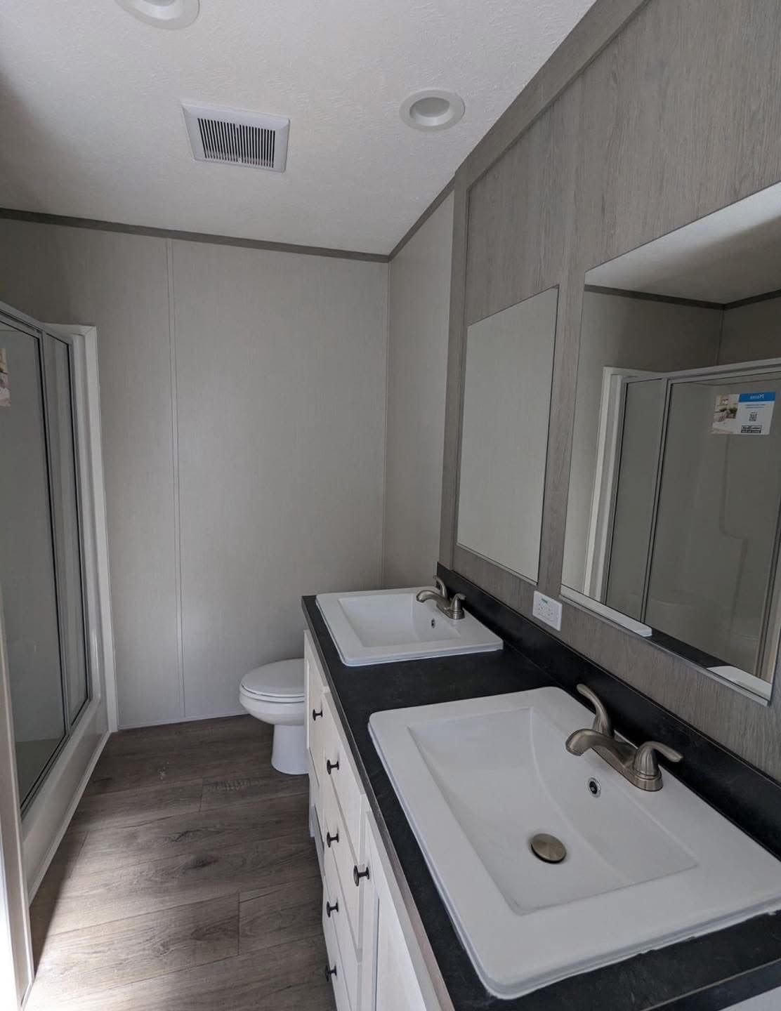 Modern bathroom with dual sinks on a dark countertop, light wood cabinets, large mirrors, a toilet, and a glass-enclosed shower. Bright and clean ambiance.