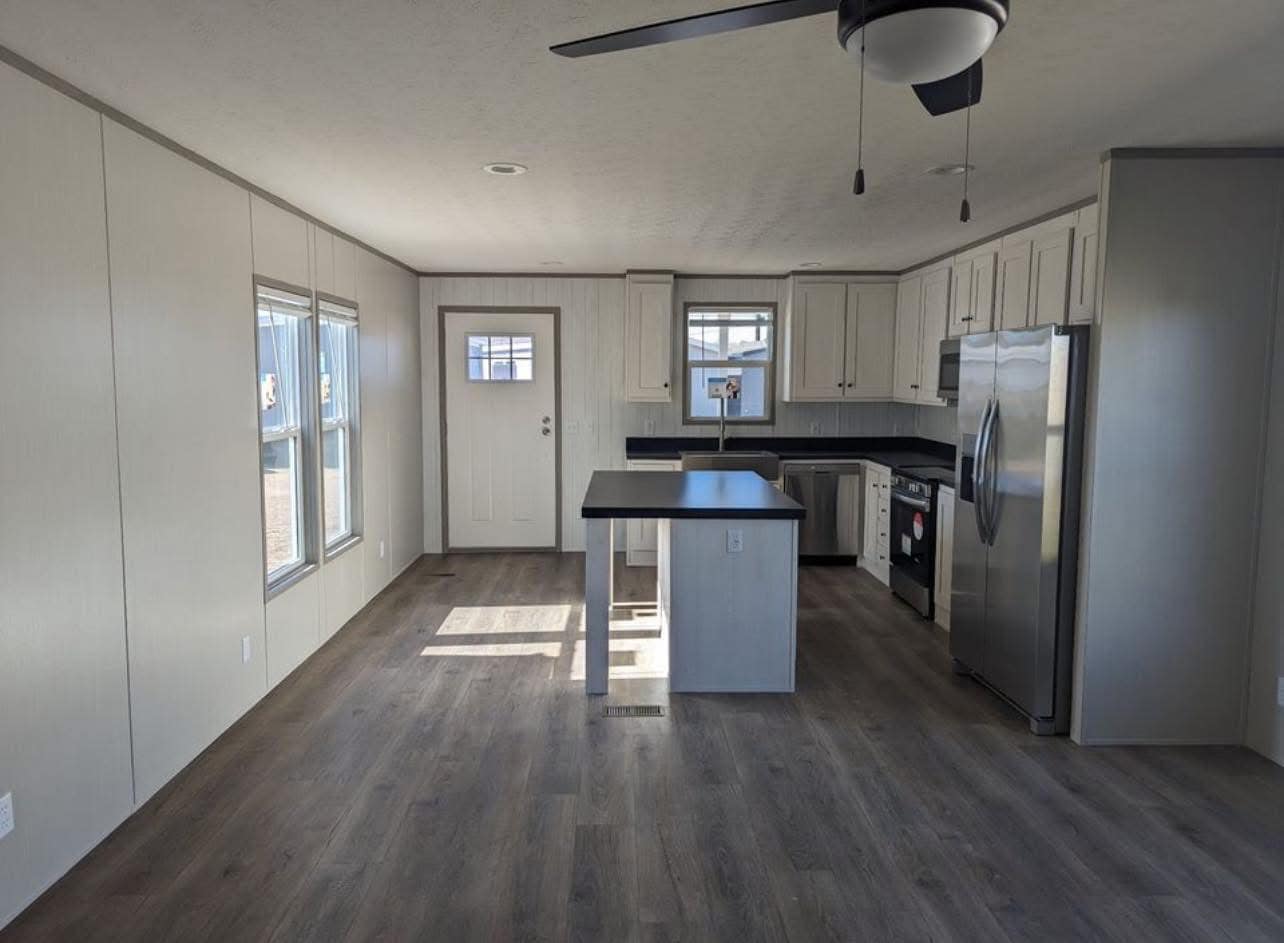 Modern kitchen with light gray cabinetry, black countertops, and stainless steel appliances. A central island and ceiling fan enhance the airy space.