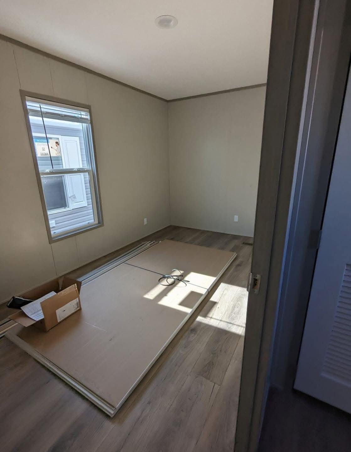 Empty room with beige walls and wooden floor. Sunlight streams through a window, illuminating a cardboard box and wooden panels on the floor, suggesting ongoing renovation.