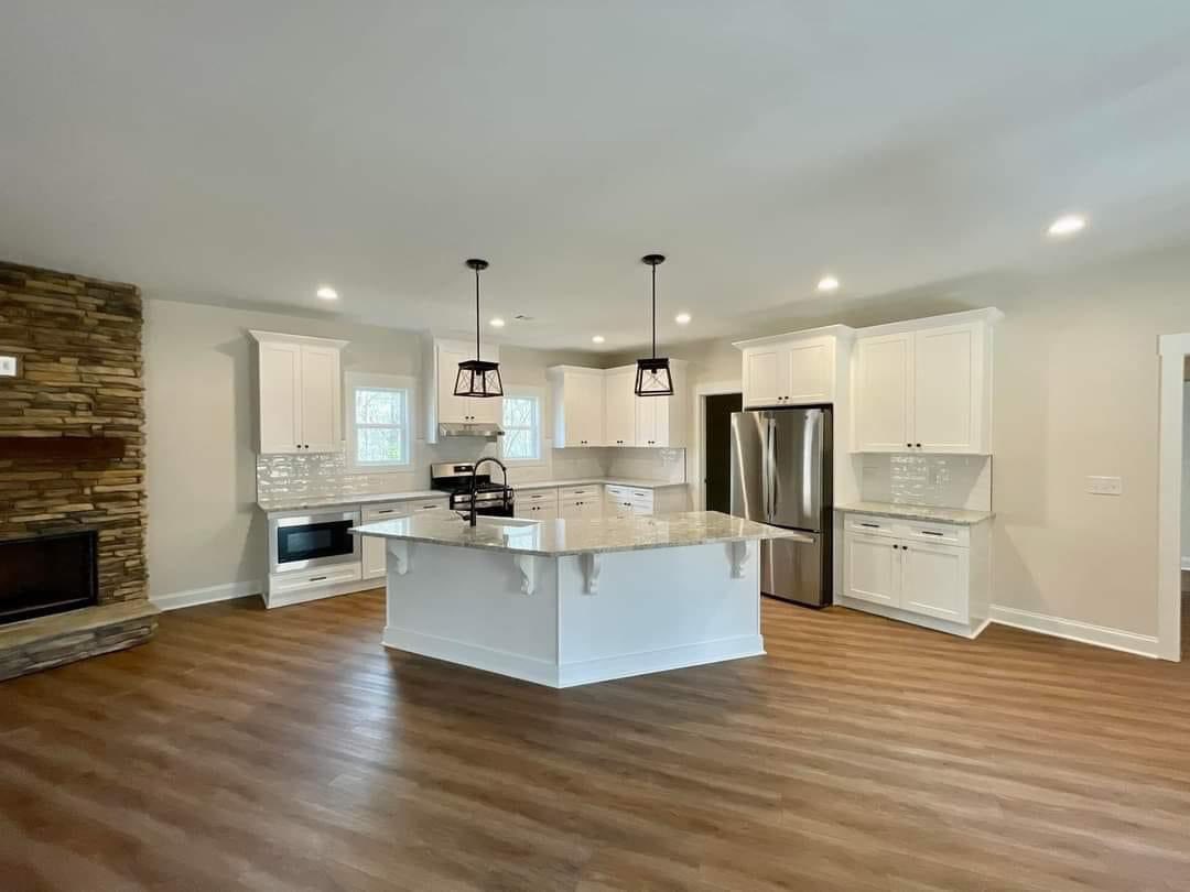 Modern kitchen with white cabinets, granite island, and stainless steel appliances. Wooden floor and a stone fireplace create a warm, inviting atmosphere.
