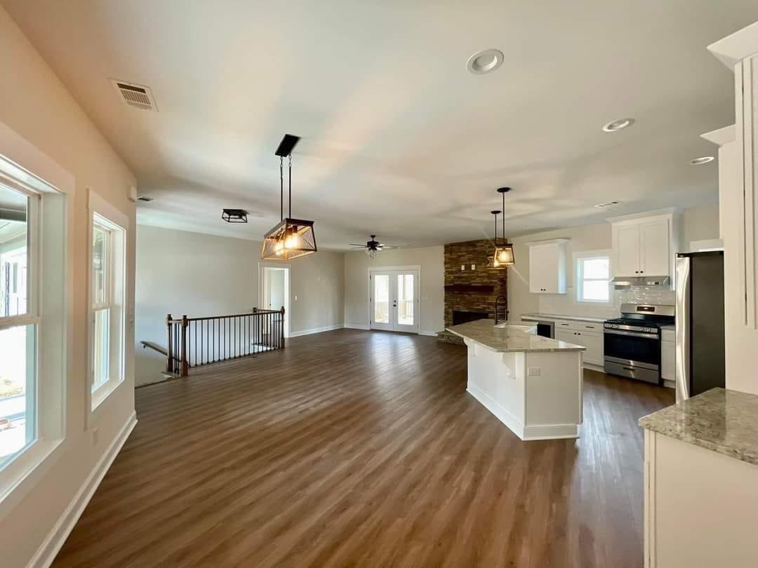 Spacious open-concept living area with light wood flooring, large kitchen island, and white cabinets. Stone fireplace and modern lighting evoke warmth.