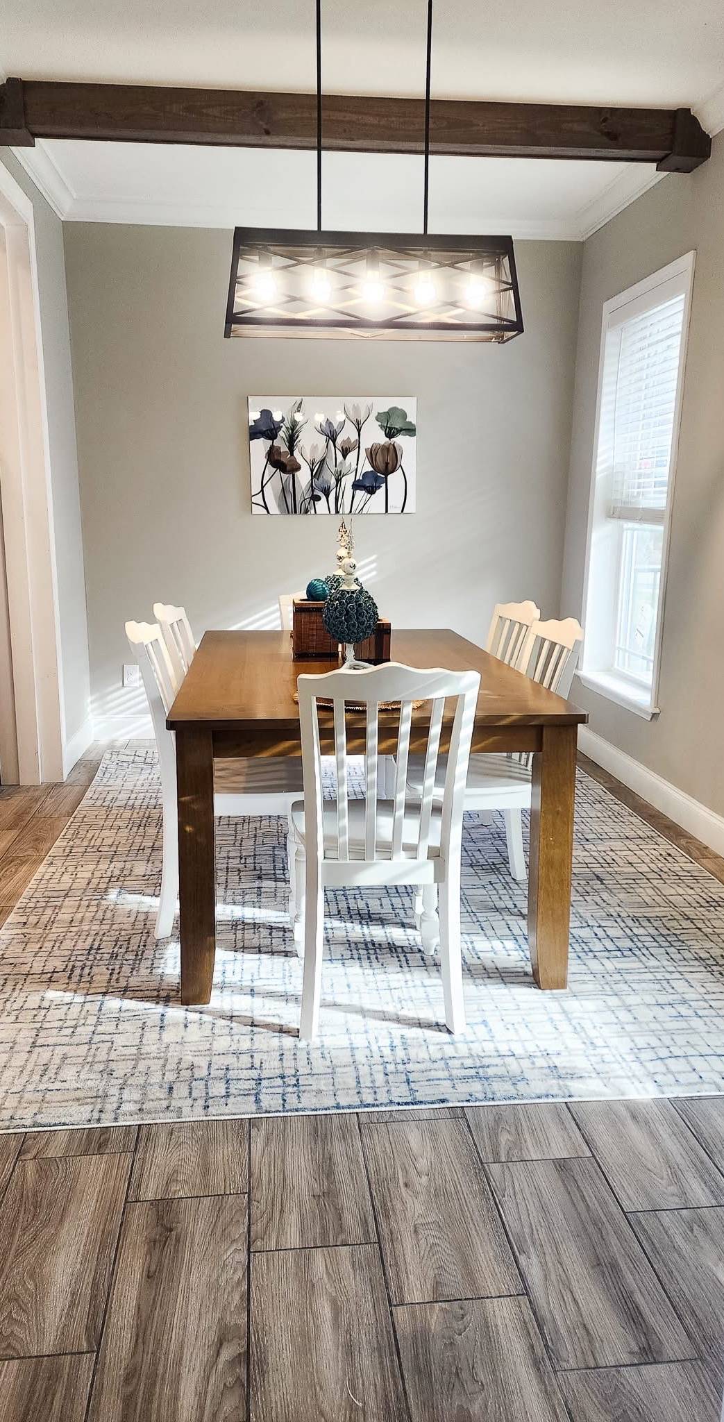 A dining room with a wooden table and four white chairs on a patterned rug. A modern light fixture hangs above, and artwork of flowers decorates the wall.