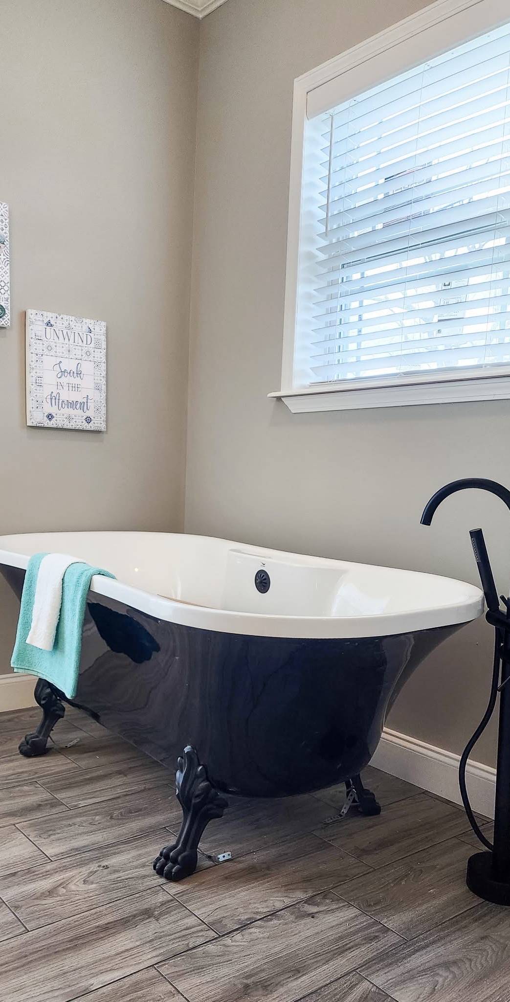 Elegant bathroom with a classic black clawfoot bathtub centered, draped with a teal towel. Neutral walls, wooden floor, and a window add a serene ambiance.