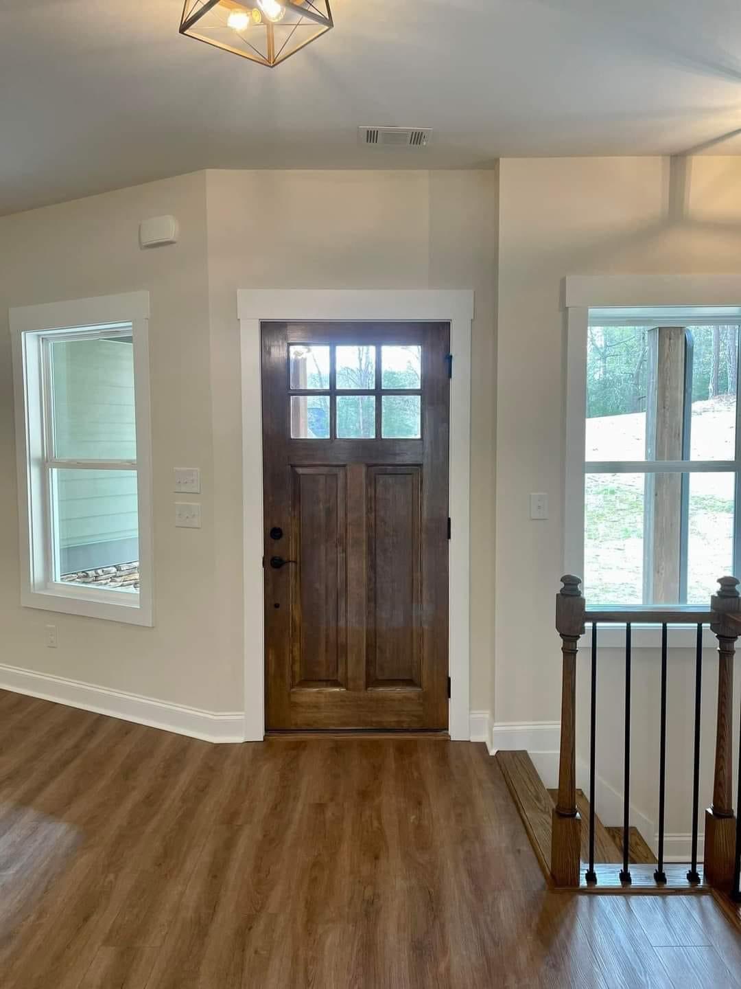 Wooden front door with glass panes, flanked by large windows and leading to a staircase. Warm light fills the room, emphasizing the wooden floor.
