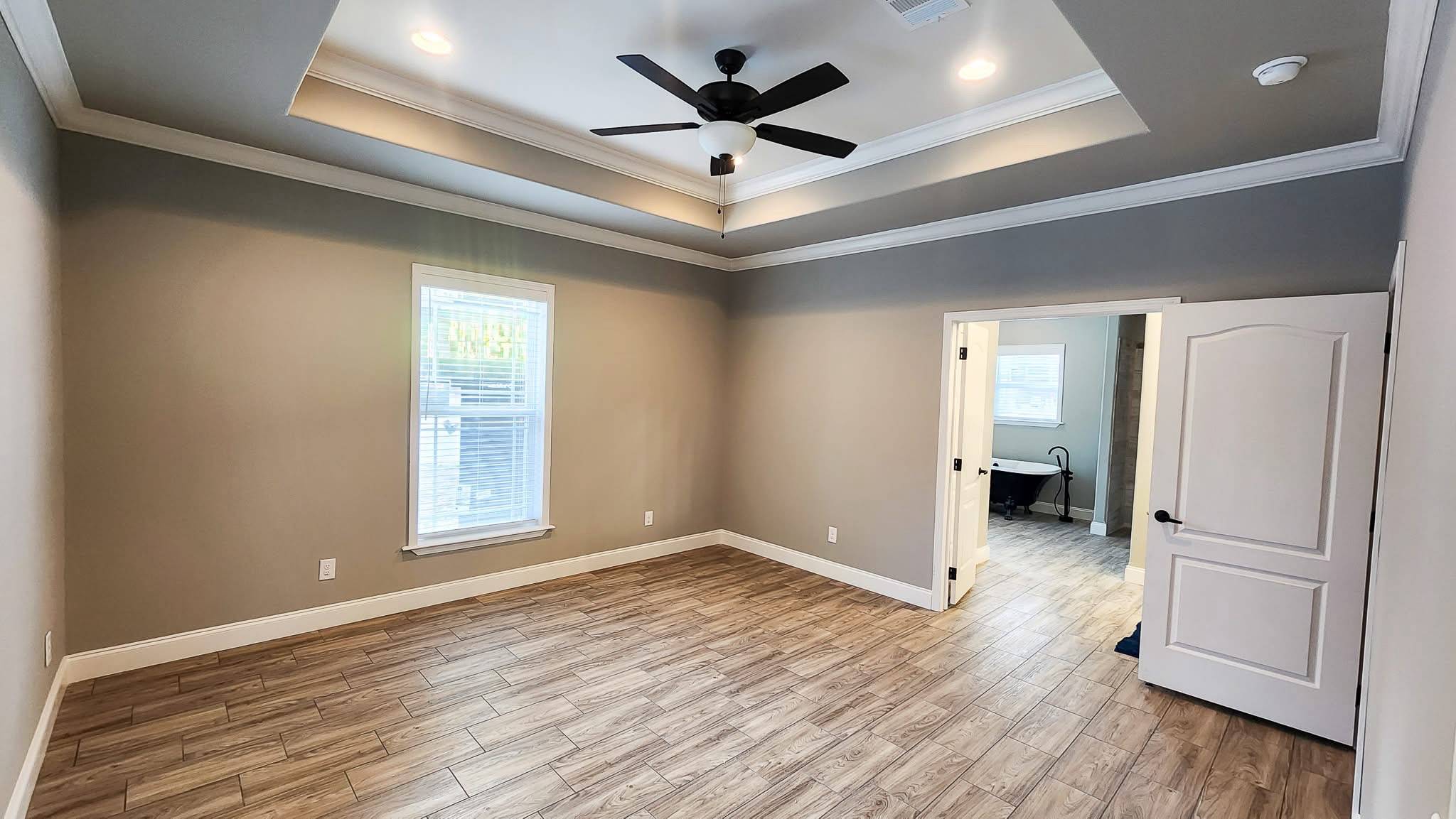 A spacious, empty room with taupe walls and wood flooring. Features a tray ceiling with recessed lighting and a black ceiling fan. An open door leads to another room.