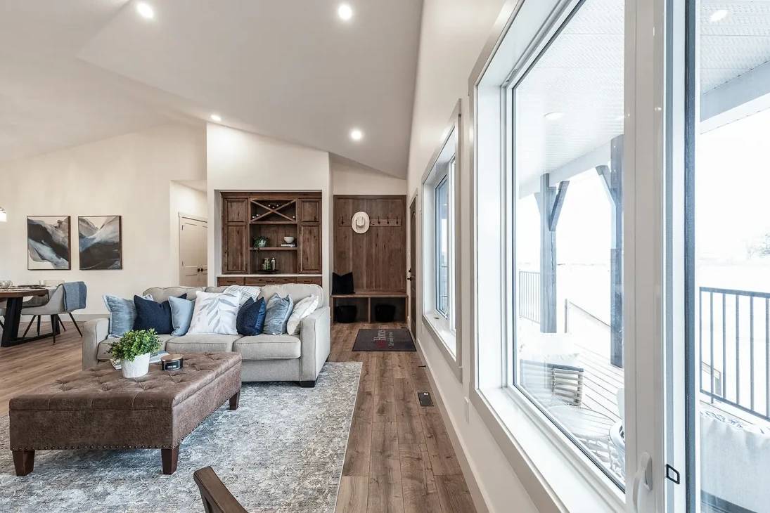 A modern living room with a cozy beige sofa adorned with blue and white cushions. A rustic wooden shelf is on the back wall. Large windows on the right allow natural light, and the atmosphere feels inviting and serene. A soft area rug and upholstered ottoman enhance comfort.