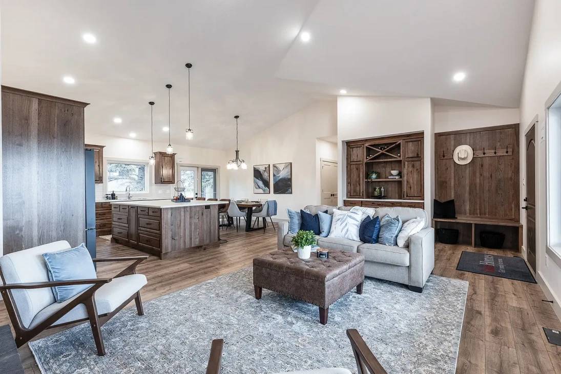 A cozy open-plan living area with wood accents features a gray sofa, blue pillows, and a plush ottoman. A kitchen island, dining table, and warm lighting complete the space.