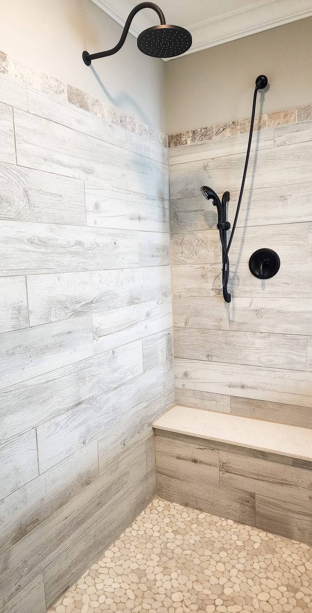 A modern shower features light wood-textured tiles on walls and bench, pebble-patterned floor, and a black rain showerhead, creating a serene ambiance.