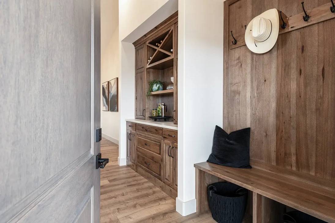Warm, rustic entryway with wooden cabinetry, wall hooks holding a white hat, and a bench with a black pillow. Natural, inviting atmosphere.