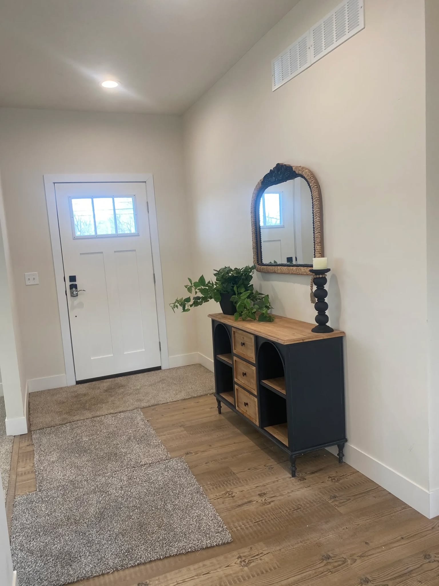 A bright entryway with a wooden floor and soft carpeting features a classic black console table adorned with a leafy plant and candle, positioned under a large mirror. The white front door with square window panes adds a welcoming touch.