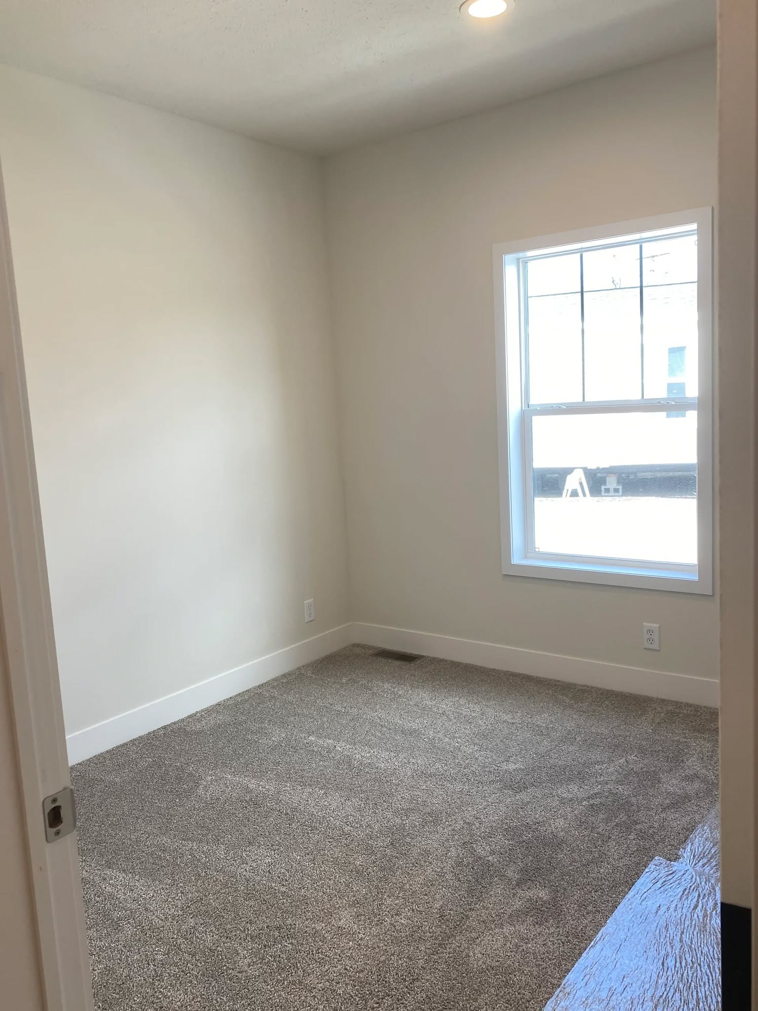 A small, empty room with beige walls and a plush carpeted floor. A single window allows natural light, creating a calm, inviting atmosphere.