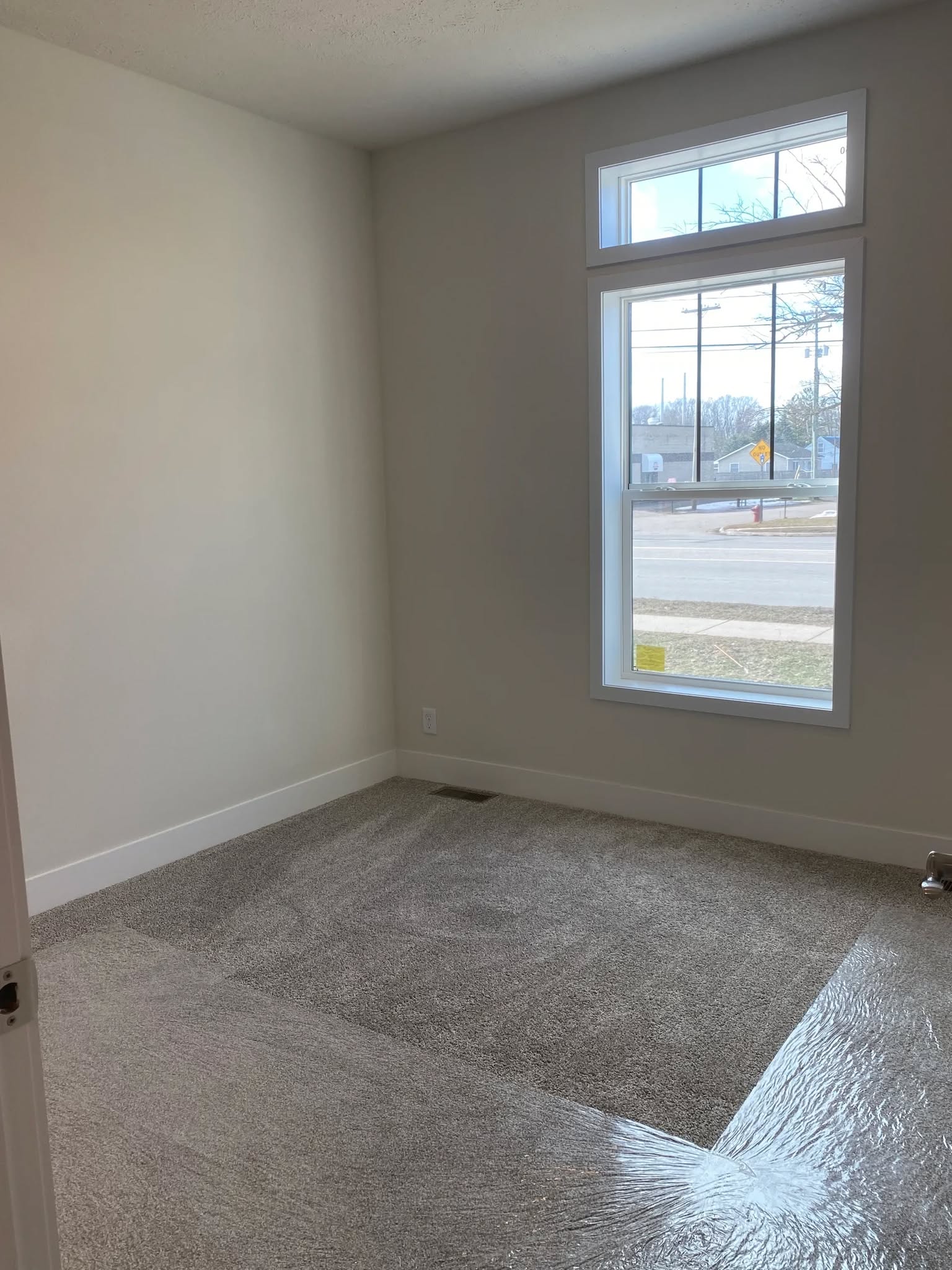 Empty room with light gray walls and carpet, a large window with two sections, and a view of a street outside. Bright, minimalist, and serene.